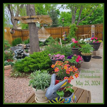 Assorted planters blooming among fenced in backyard trees, shrubs, water fountain and hammock.