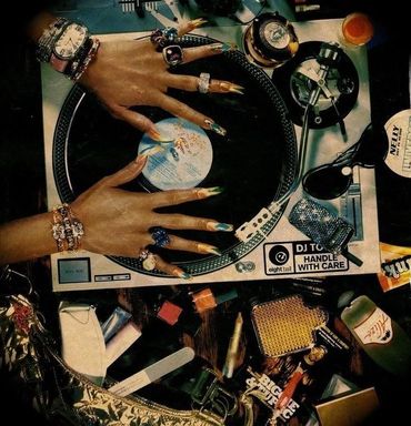 Hands with long nails and jewelry on a DJ turntable surrounded by accessories.