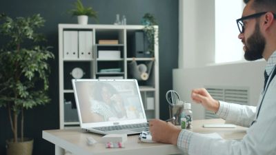 A doctor conducts a video consultation with a patient using a laptop in a medical office.