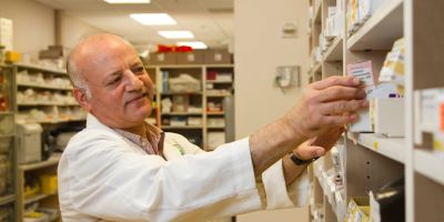 A pharmacist in a white coat selects a medication box from a pharmacy shelf.