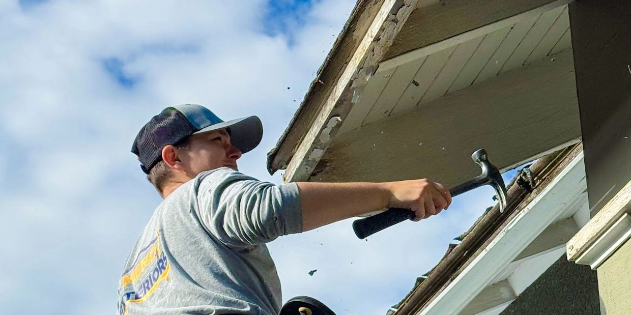 Man removing old wood from roof edge with a hammer.