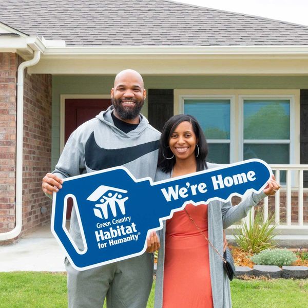 Couple holding a 'We're Home' key sign in front of their new house.