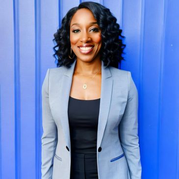Smiling woman in a gray blazer stands against a vibrant blue wall.