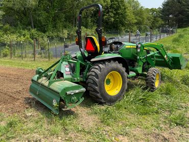 Green John Deere tractor working on a farm field with trees in the background.