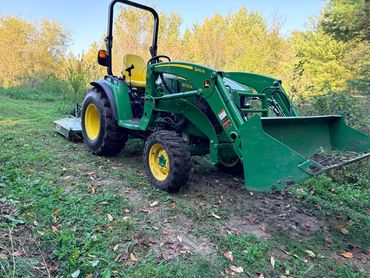 Green John Deere tractor with front loader in a wooded area.