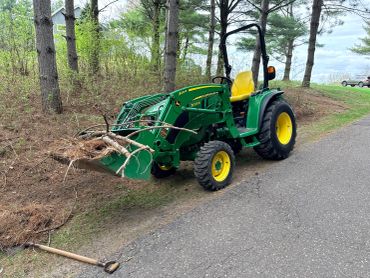 John Deere tractor with front loader holding branches on a wooded roadside.
