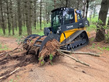 A Deere 325G tracked skid steer clearing tree roots and branches in a wooded area.