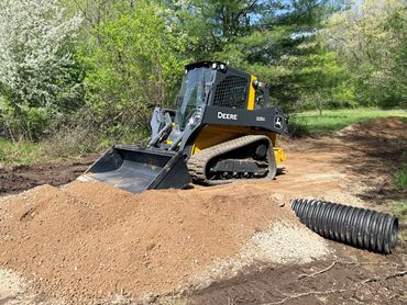 John Deere 325G skid steer loader working on soil near a drainage pipe in a wooded area.
