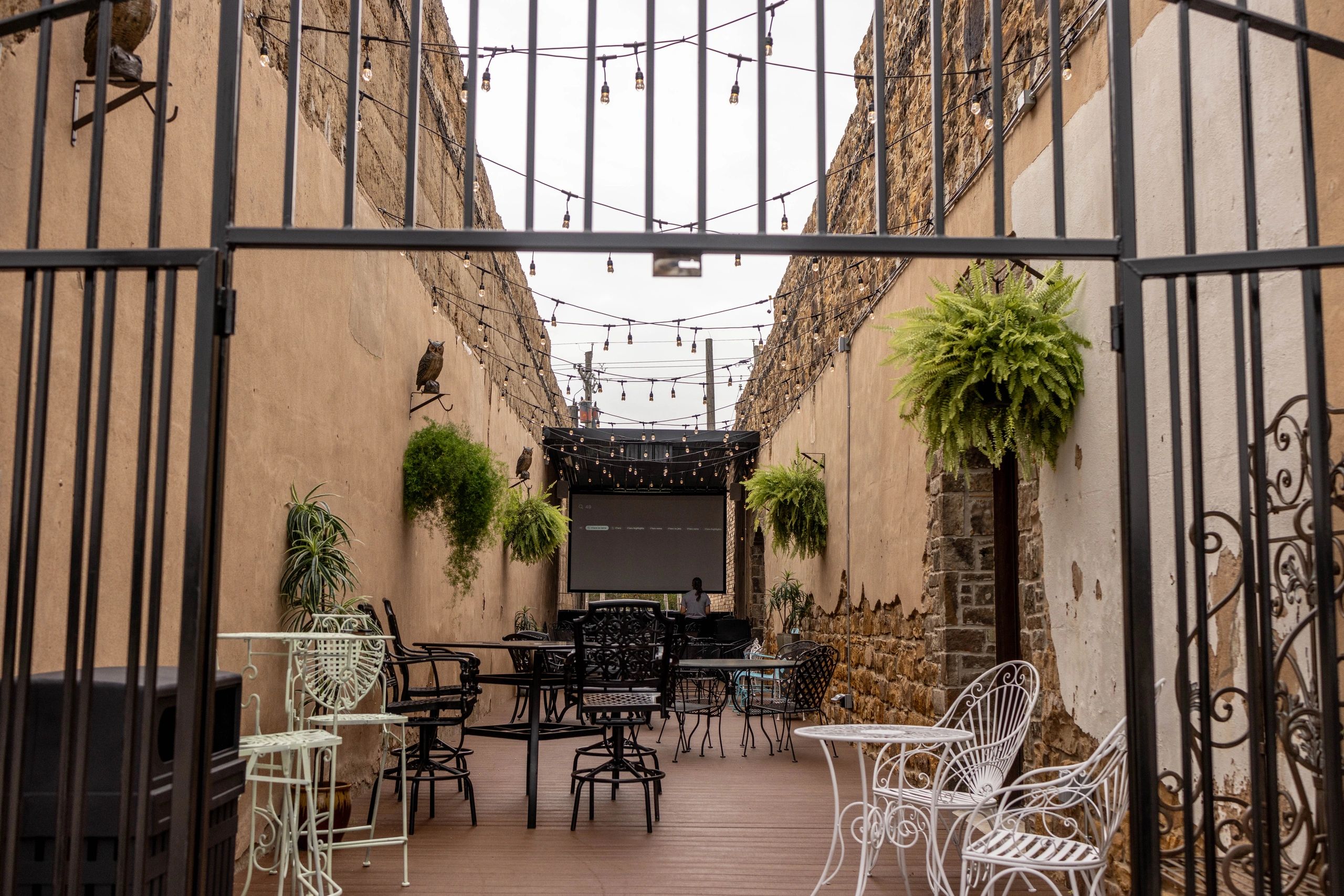 Outdoor patio with string lights, hanging plants, and wrought iron furniture, viewed through a gate.