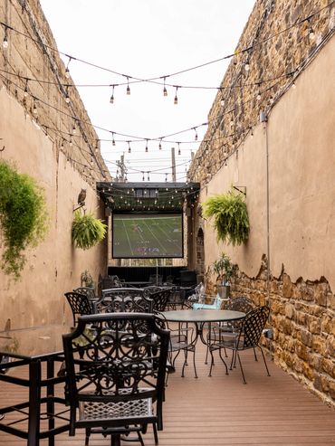 Cozy outdoor patio with string lights, metal chairs, and a large screen showing a football game.