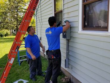 Two electricians working on an outdoor electrical panel beside a house.