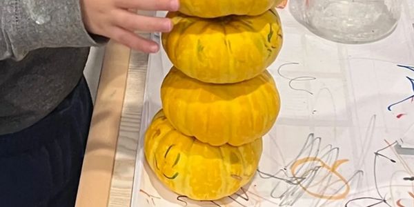 Four small yellow pumpkins stacked on a table with a child's hand nearby.