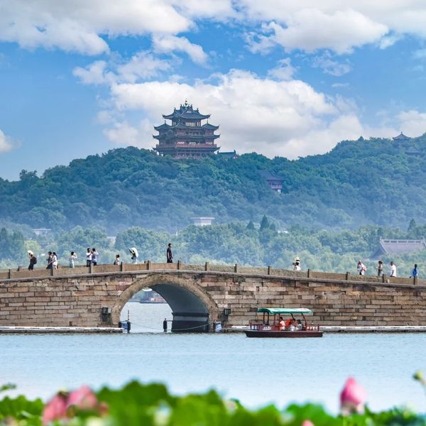 Scenic view of a stone bridge with people, a boat on water, and a traditional pagoda on a hill.