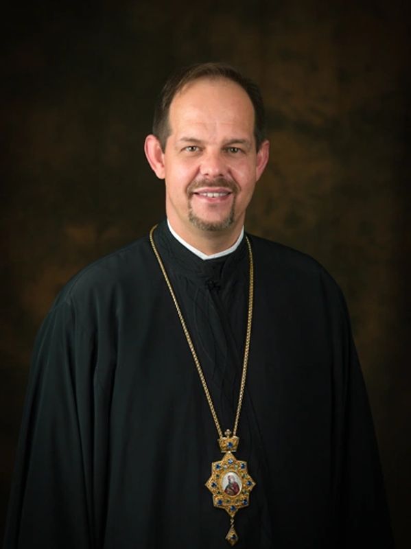 Clergyman in black robe with ornate religious pendant smiling.