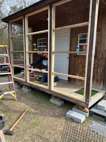 Man building a wooden and wire frame structure attached to a small wooden porch.