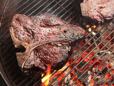 Two thick T-bone steaks grilling over hot charcoal flames.