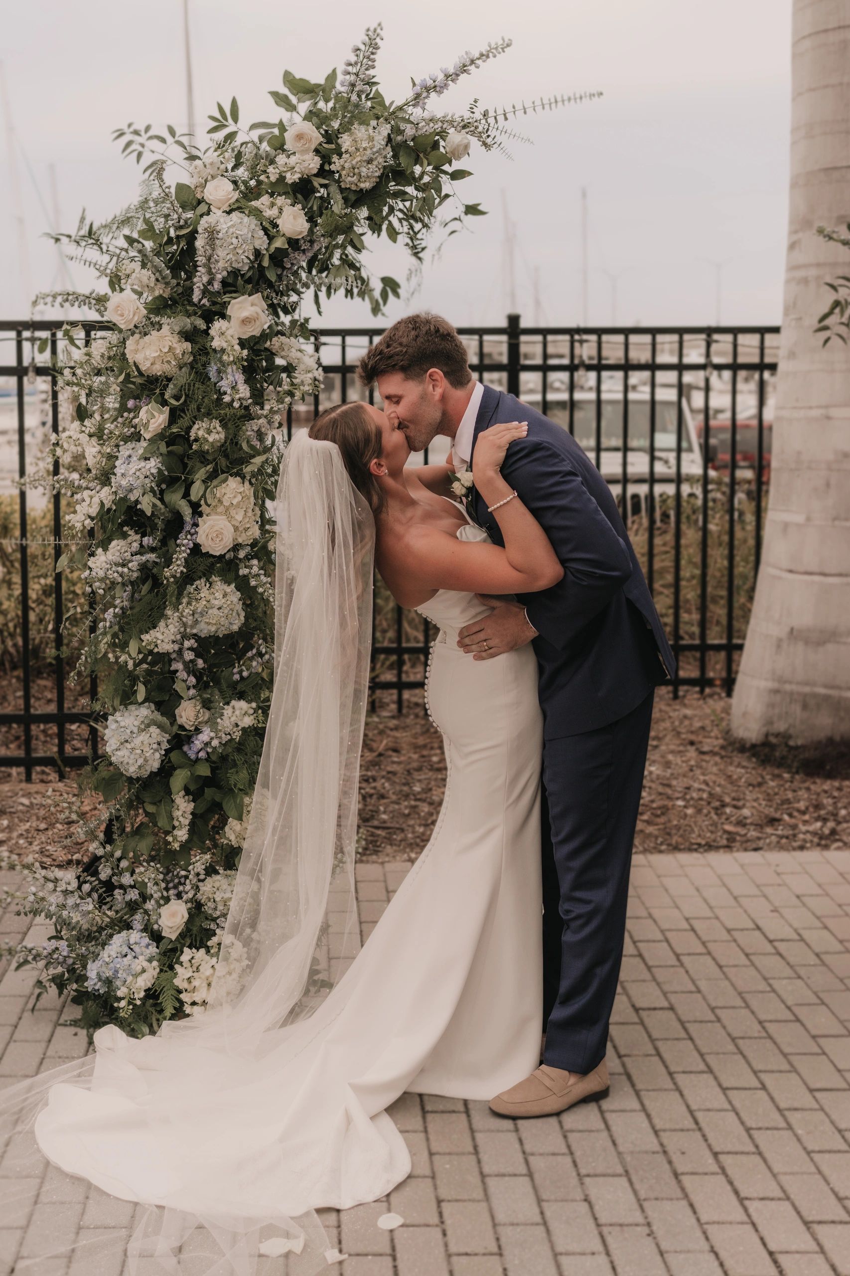 Newlyweds share a romantic kiss under a floral wedding arch outdoors.
