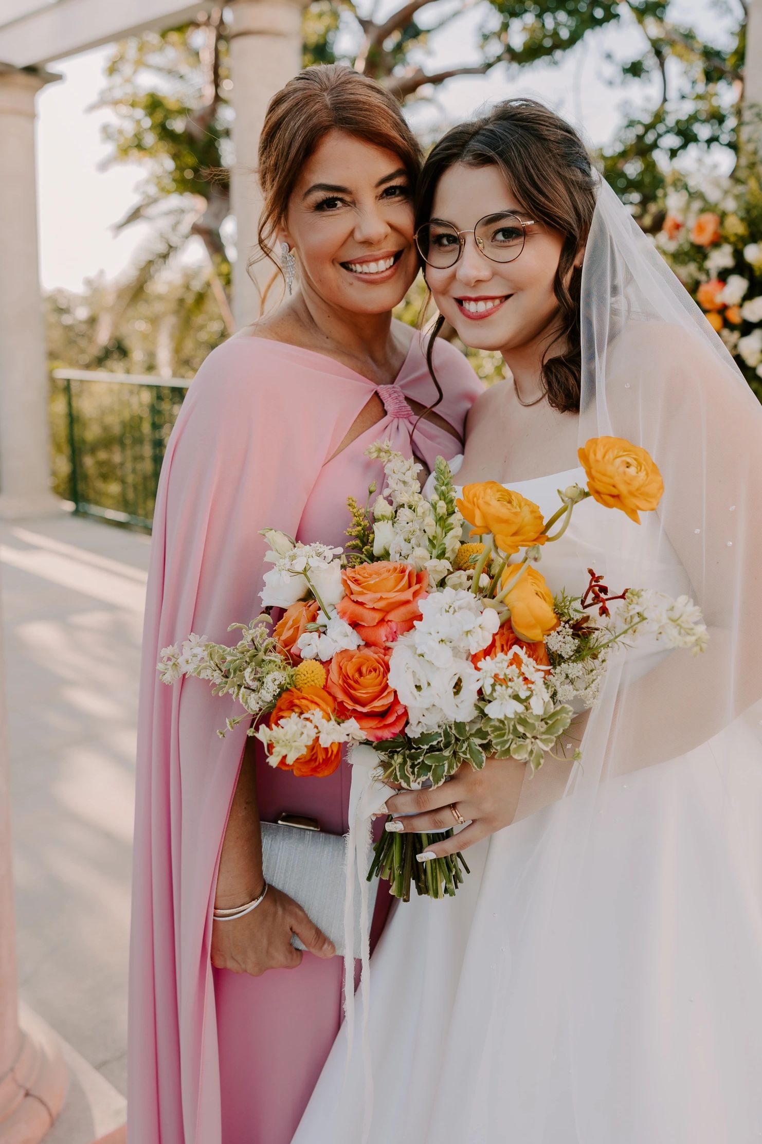 Bride and woman in pink dress smiling with a bouquet of flowers outdoors.