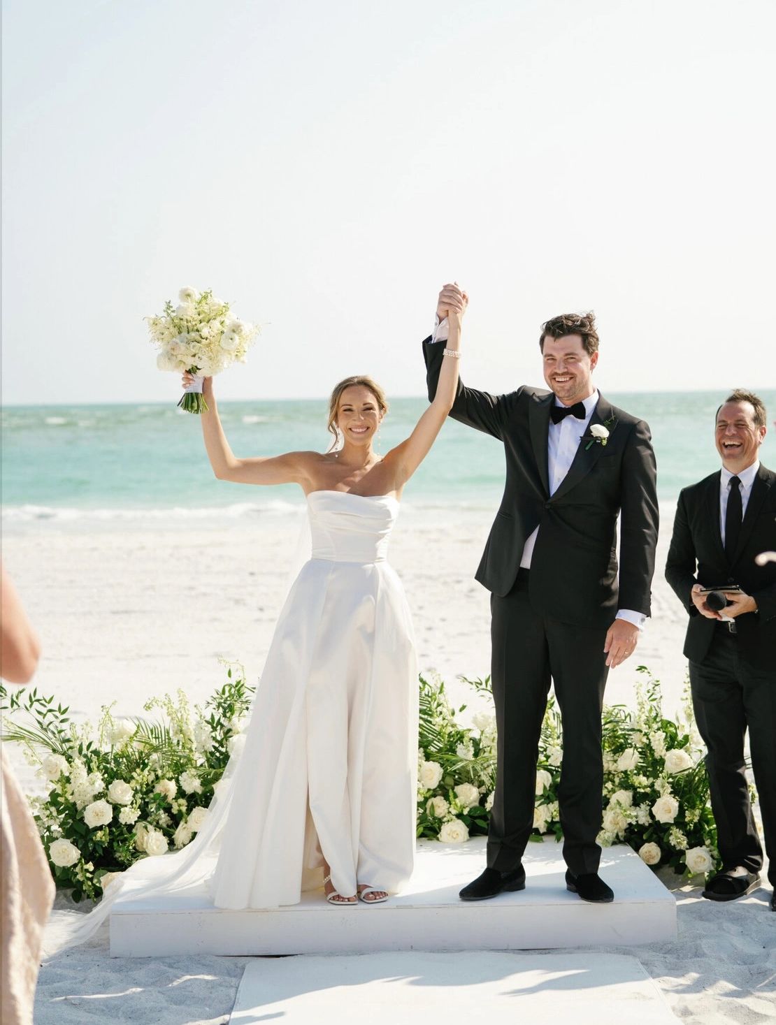Bride and groom celebrate their wedding on a beach with smiles and raised hands.