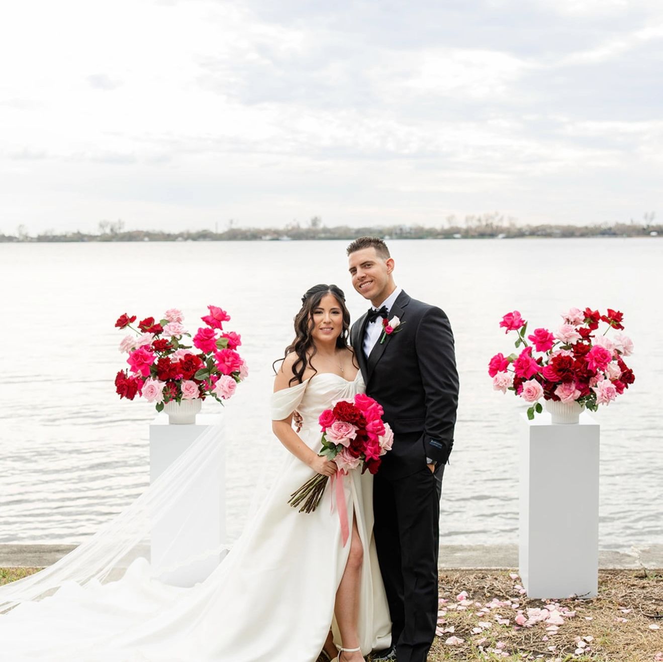 Bride and groom posing by the water with vibrant floral arrangements.