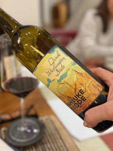 A hand holding a wine bottle labeled "Drink Washington State" with a glass of red wine in the background.