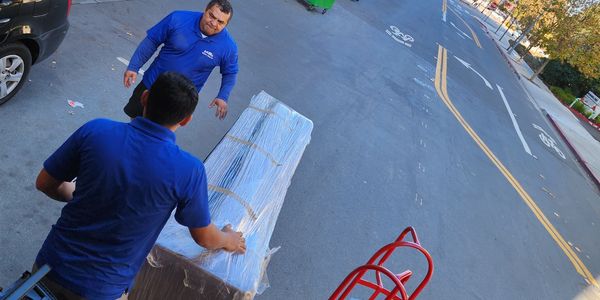 Two men in blue shirts unloading a large wrapped item from a truck onto the street.
