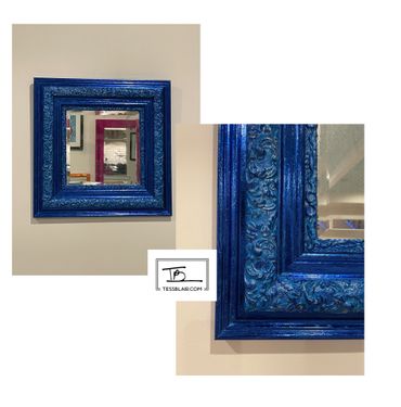 Ornate blue square mirror frame with intricate floral patterns hanging on a beige wall.