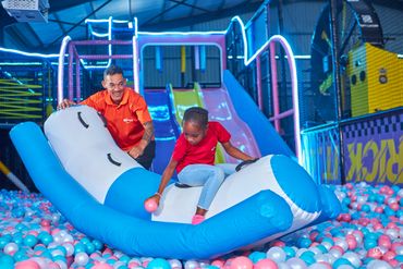 A man and a girl play on an inflatable toy in a colorful ball pit at an indoor playground.