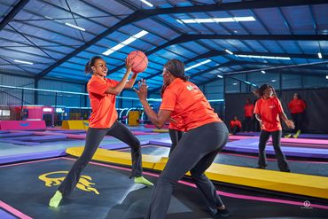 Women in orange shirts playing basketball on trampolines indoors.