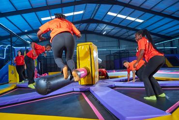 Group of people playing on a trampoline with a rotating padded beam indoors.