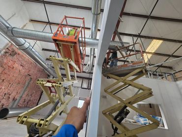Two workers on scissor lifts installing ductwork in a spacious industrial building.