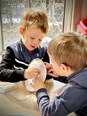 Two boys working together on a craft project involving a paper plate and string.