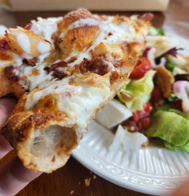 Hand holding a cheesy pizza slice with a salad plate in the background.