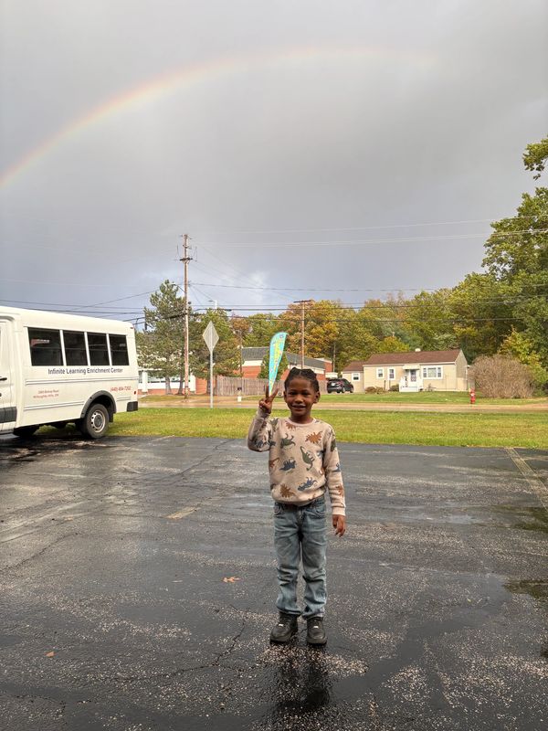 A child stands on wet pavement under a rainbow, smiling and holding up a peace sign.