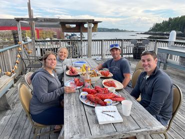 two couples sitting at a picnic table on a dock about to eat a lobster dinner