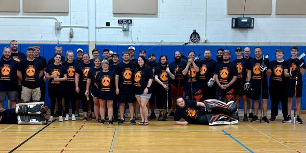 Large group in matching shirts posing in gym with hockey sticks and goalie gear.