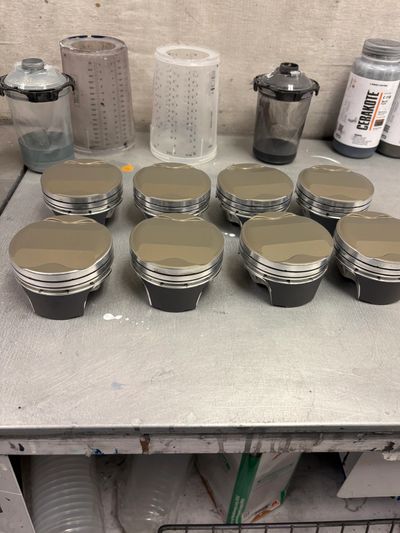 Eight metallic pistons with a smooth finish arranged on a workbench.