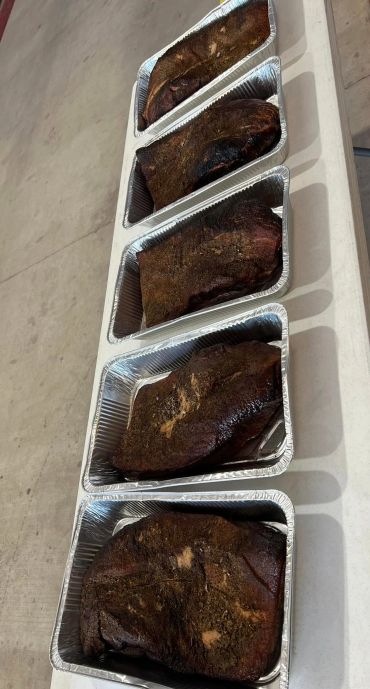 Five trays of smoked or grilled meat on a white table.
