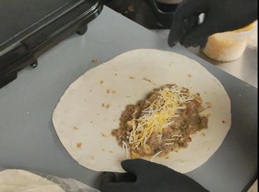 Gloved hands preparing a burrito with meat and cheese.