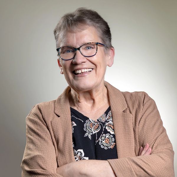 Smiling elderly woman with glasses and crossed arms wearing a patterned blouse and brown jacket.