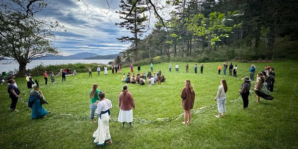 People forming a large circle outdoors on a grassy field near water and trees.