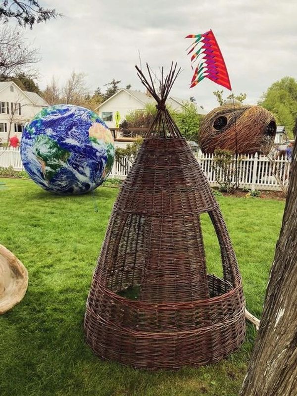 Wicker tepee structure on grass with Earth and coconut-shaped sculptures in background.