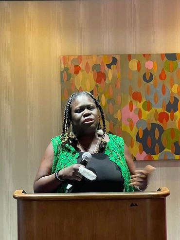 Woman speaking at a podium with a colorful abstract painting in the background.