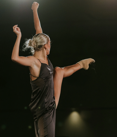 A dancer in a black dress performs a high leg lift on stage.