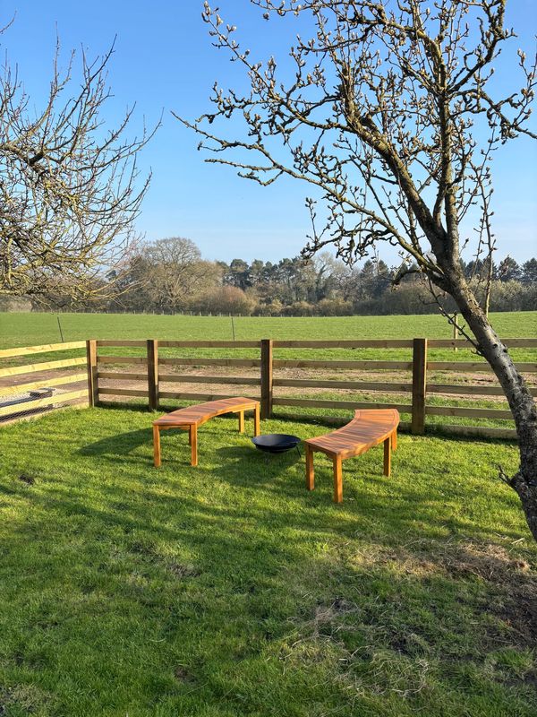 Curved wooden benches and a fire pit in a fenced grassy yard under a clear blue sky.