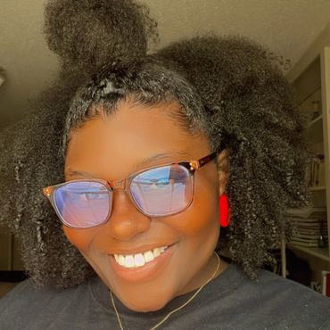 Smiling woman with glasses and voluminous natural hair wearing a black shirt with red lettering.