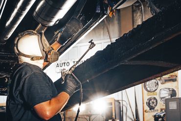Worker wearing protective gear uses a blowtorch on a vehicle's underside.