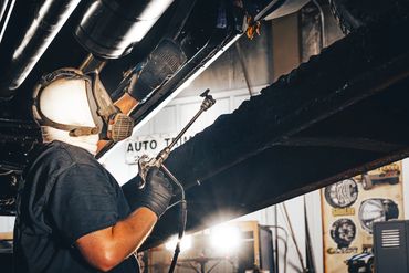 Worker wearing protective facial gear uses a high pressure rustproof spray on a vehicle's underside.