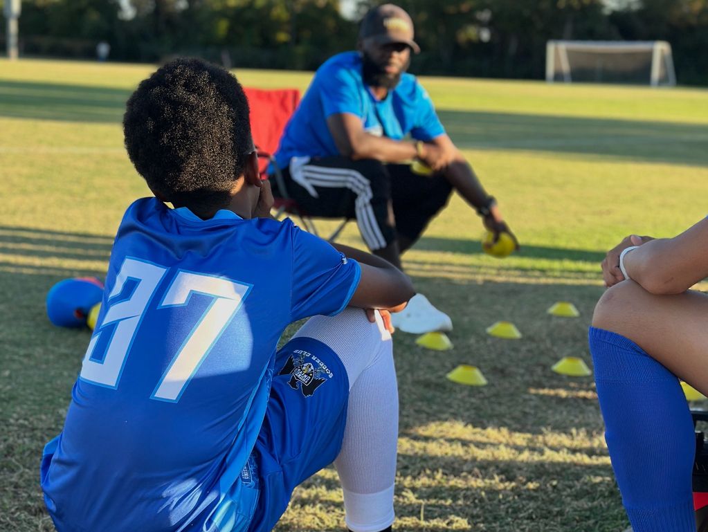 Young soccer players listen to their coach during a practice session on a sunny field.