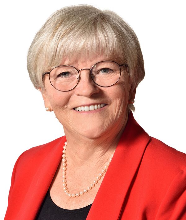 Smiling elderly woman in glasses and red blazer with pearl necklace.